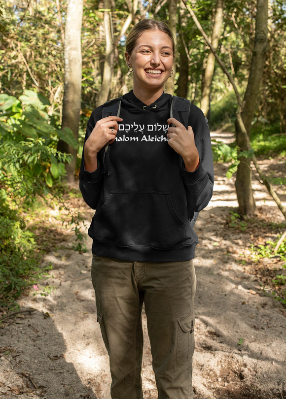 Smiling woman wearing a black hoodie with the words Shalom Aleichem in English and Hebrew. Walks along a hiking trail wearing a backpack. 